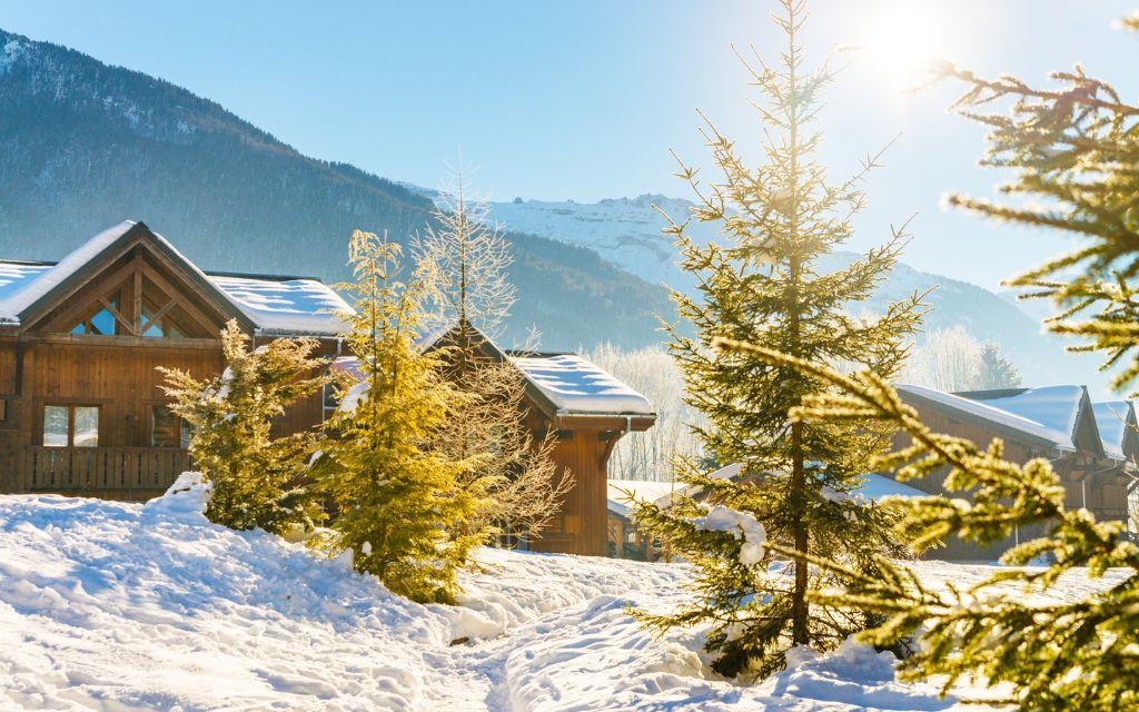 Slide Chalets en bois sous la neige aux Fermes de Samoëns by Ateya, avec vue sur les montagnes ensoleillées.