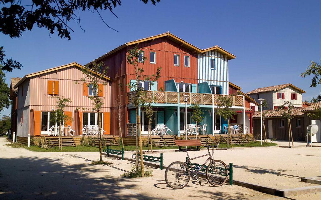 Slide Wood-clad, colourful buildings arranged in a row, with a pétanque court and bicycle rack at the Les Rives Marines residence in Le Teich